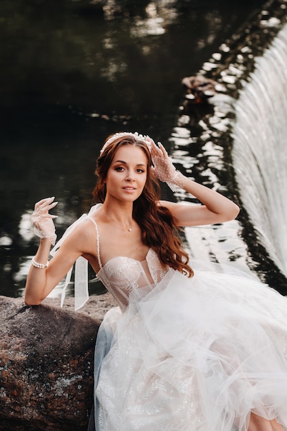 Un'elegante sposa in abito bianco, guanti e piedi nudi è seduta vicino a una cascata nel parco godendosi la natura. un modello in abito da sposa e guanti in un parco naturale. bielorussia