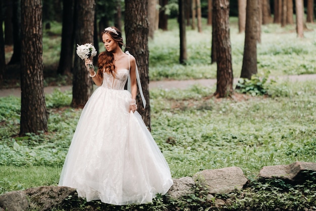 An elegant bride in a white dress and gloves holding a bouquet stands by a stream in the forest, enjoying nature.A model in a wedding dress and gloves in a nature Park.Belarus