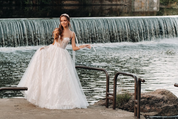 Элегантная невеста в белом платье и перчатках стоит у реки в парке, любуясь природой. Модель в свадебном платье и перчатках в природном парке. Беларусь