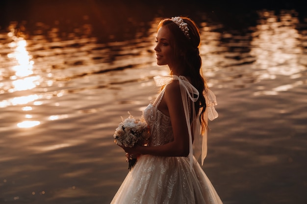 An elegant bride in a white dress and gloves stands by the river in the Park with a bouquet, enjoying nature at sunset.A model in a wedding dress and gloves in a nature Park.Belarus