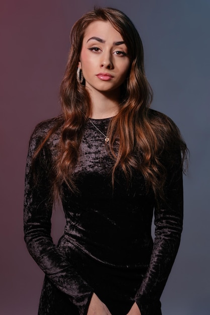Elegant brunette woman in stylish jewelry and black velvet dress posing against colorful background ...