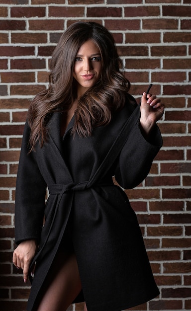 Elegant brunette woman with long curly hair wearing coat, posing near brick wall