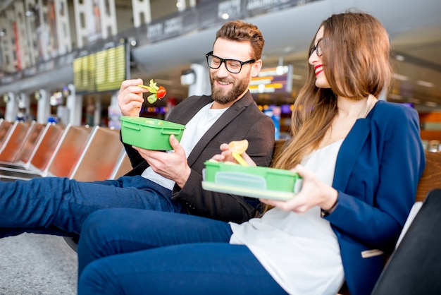 Elegant business couple eating with lunch boxes sitting at the waiting hall in the airport. Having a snack during business trip