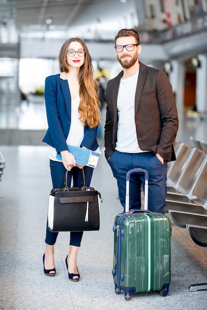 空港の待合室でスーツケースと飛行機のチケットを持って立っているエレガントなビジネスカップル。出張の概念