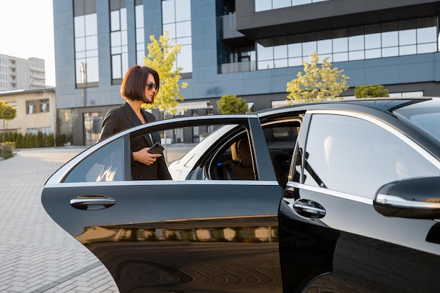 Элегантная деловая женщина садится в роскошный черный автомобиль возле офисного здания или отеля Концепция деловых перевозок и путешествий
