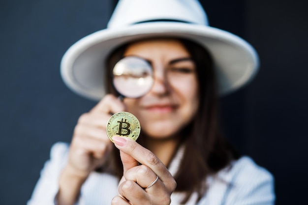 Elegant business woman in a white hat holds a magnifying glass and looks at bitcoin The concept of research and study of the cryptocurrency market