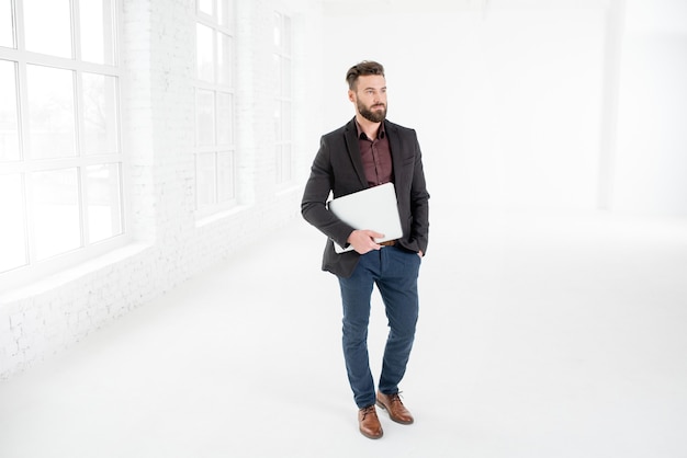 Elegant businessman in the dark suit standing with laptop in the white office interior