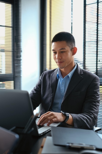 Elegant businessman looking at laptop screen reading financial report online and writing important notes