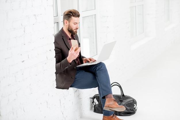 Elegant businessman sitting on the window with laptop and mobile phone in the white office interior