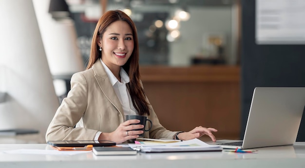 Elegant businesswoman holding mug and working on laptop computer at office smiling and looking at camera