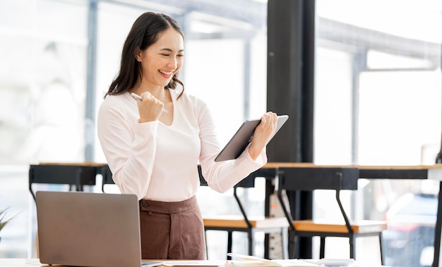 Elegante donna d'affari seduta in ufficio con tavoletta digitale eccitata donna d'affari asiatica che alza le mani per congratularsi mentre lavora al computer portatile in ufficiox9