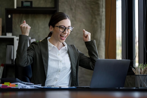 Elegant businesswoman sitting in office with laptop Excited businesswoman raising hands to congratulate while working on laptop in office