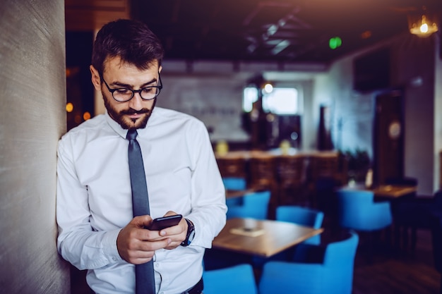 Elegant caucasian bearded handsome businessman in shirt, tie and with eyeglasses leaning on wall in cafe and using smart phone for reading or sending message.