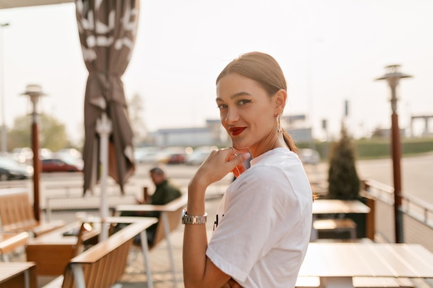 Elegante donna affascinante con capelli scuri raccolti con labbra rosse che indossa una maglietta bianca in posa alla luce del sole ritratto bella ragazza con i capelli lunghi che fa colazione all'aperto