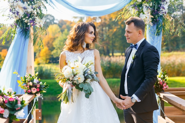 Elegant curly bride and happy groom outdoors on the background the lake. Creative stylish wedding ceremony