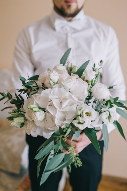 Elegant delicaat boeket van de bruid gemaakt van witte pioenrozen, hortensia's, rozen en een tak groen in de handen van de bruidegom in de kamer.