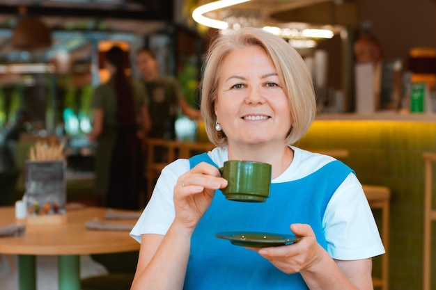 Elegant elderly senior woman in a blue sundress holding a cup in her hands sitting in a cozy cafe and relaxing enjoying