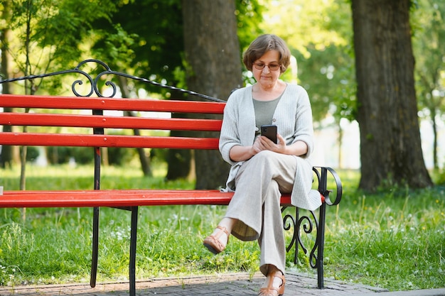 Elegant elderly woman in the shirt is sitting on the bench in a park on a warm day