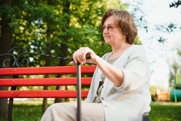 Elegant elderly woman in the shirt is sitting on the bench in a park on a warm day