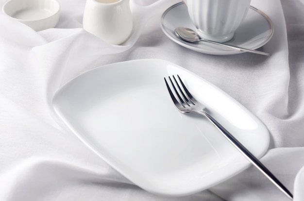 Elegant empty plate, cutlery, cup of coffee on sunny morning, clean white tablecloth background, 