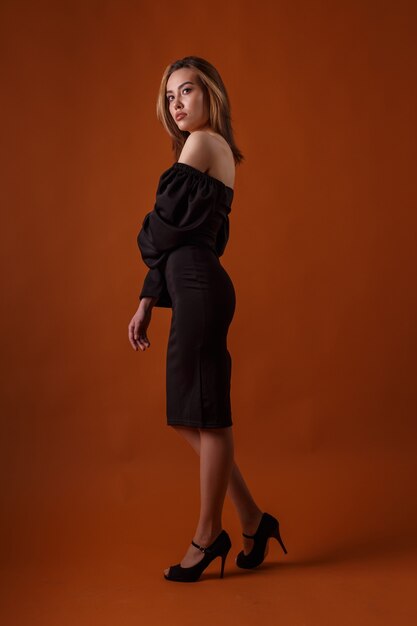 Elegant fashion model in black dress with deep neckline, high heel shoes posing on orange background