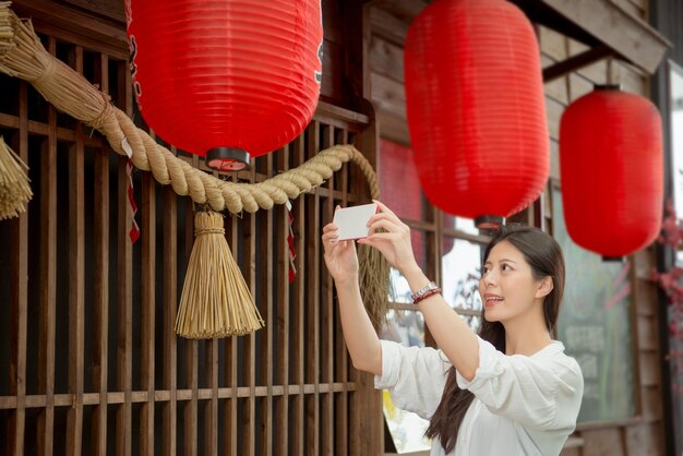 Elegant female tourist holding mobile phone camera app to\
taking traditional japanese tea lanterns decorated pictures with\
japanese text meaning celebration in japan season festival.