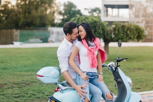 Elegant girl in casual attire smiling to boyfriend sitting with him on scooter. Outdoor portrait of strong man in white shirt spending time with wife in summer vacation.