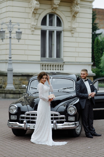 Sposa splendida elegante e sposo bello che si abbracciano in elegante automobile nera alla luce.