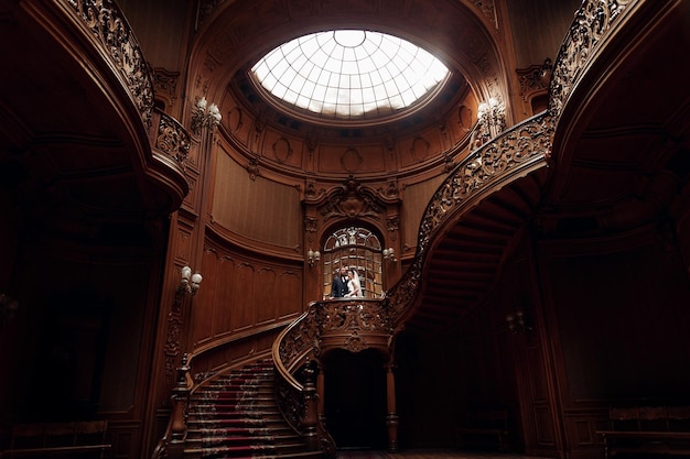 Elegant gorgeous bride and stylish groom standing on wooden stairs in amazing old rich room unusual wedding couple in retro style luxury wedding concept