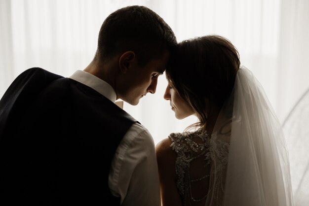 優雅な新郎と美しい花嫁が頭を下げて窓の前に立っています。結婚式、愛、関係の概念。低いキー。フルレングスのポートレート。
