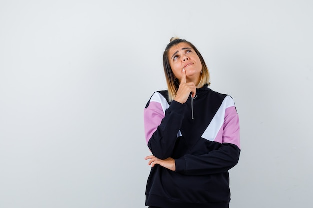 Elegant lady holding finger on chin, looking up in sweatshirt and looking thoughtful.