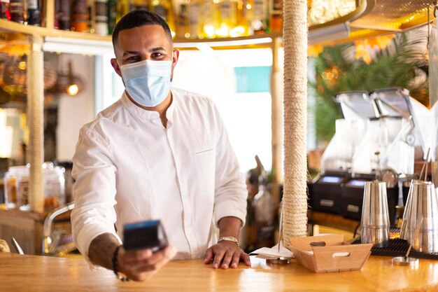 Elegant Latin American waiter with a dataphone in his hand to make a payment. He wears a medical mask. Space for text.