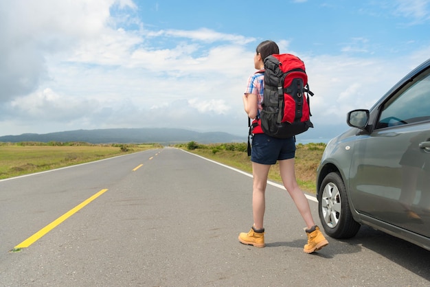 elegant leisurely female backpacker standing on asphalt road watching distant mountain scenery and parked car in roadside at summer vacation travel.