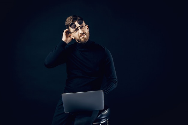 Photo elegant man in black clothes and flip-up glasses brainstorming on idea sitting with laptop.