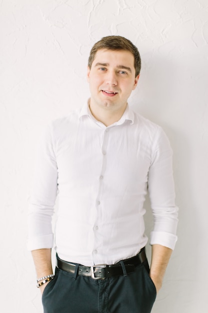 Elegant man groom in white skirt standing near white wallpaper with a hands in pockets