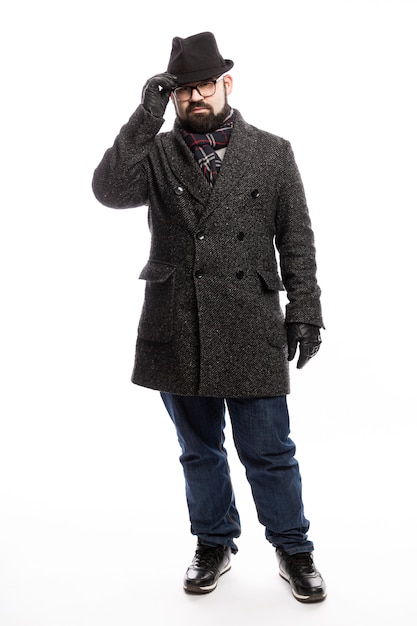 Elegant man with a beard in a coat and hat. He stands in full growth on a white wall and holds on to his hat. Vertical.