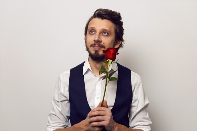 Uomo elegante con barba e occhiali il giorno di san valentino in una camicia bianca e gilet su un muro bianco sta con una rosa rossa