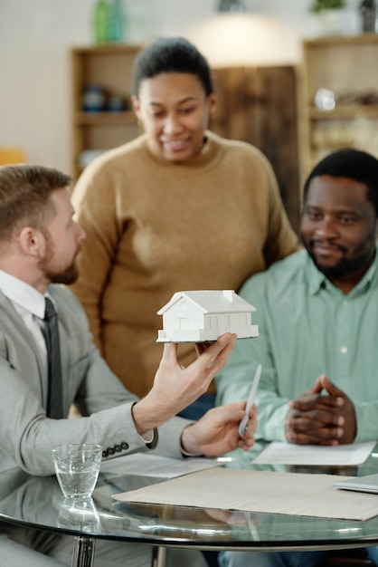 Elegant man with house model advising black couple on real estate purchase gathering at table