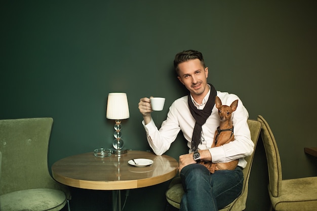 The elegant man with a little dog on hands having coffee at cozy restaurant indoors