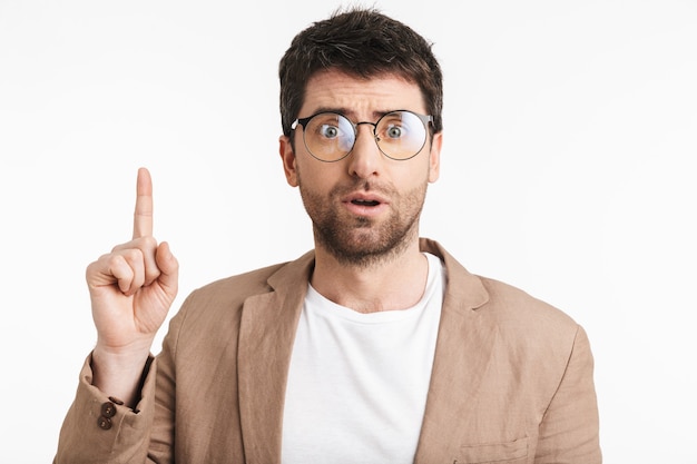 elegant man with stubble wearing jacket and eyeglasses pointing finger upward at copyspace isolated over white wall