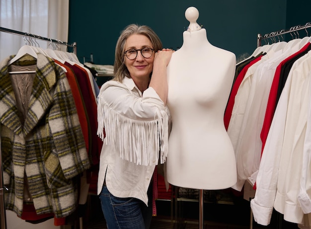 Elegant mature European woman leaning on a dressmaker's model while posing in her fashion clothing design workshop and tailoring atelier