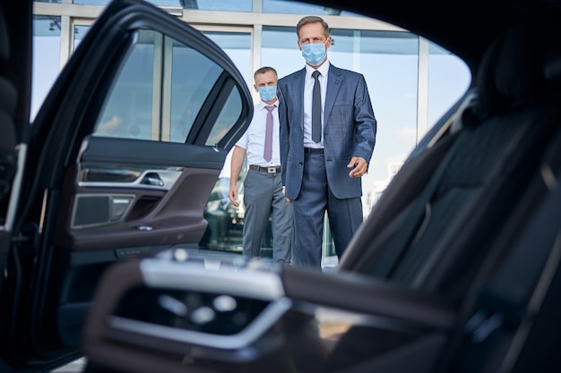 Elegant mature man in sterile mask is getting into automobile after arrival while chauffeur is holding suitcase