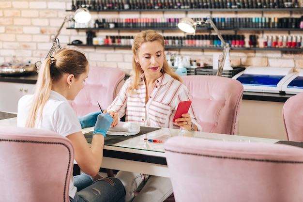 Elegant mature woman using phone and manicurist filing nails