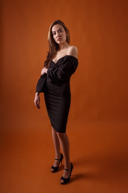 Elegant model met zwarte jurk en schoenen met hoge hakken poseren op een oranje achtergrond