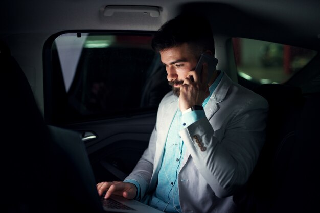 Elegant modern businessman with laptop on the back seat talking on a mobile.