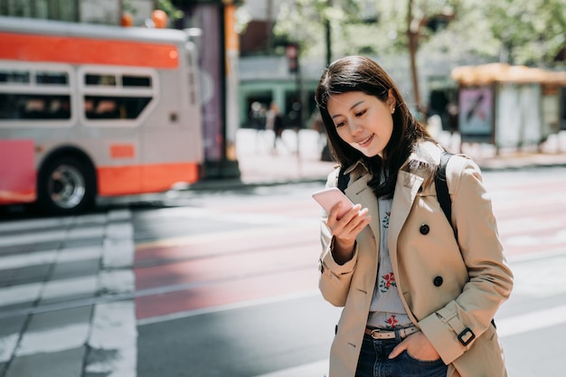 웃고 있는 친구들의 재미있는 메시지를 읽는 휴대폰을 사용하는 우아한 사무실 여성. 도로 위에 서 있는 아름다운 아시아 여성이 버스 운전 패스를 배경으로 휴대전화를 보고 있습니다. 젊은이 기술
