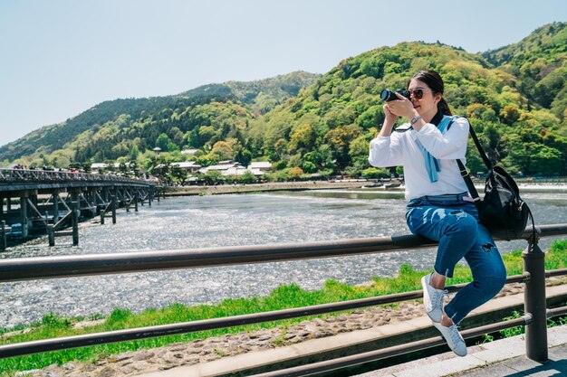 手すりに座って渡月橋を撮影する優雅な写真家。旅行レンズの男の趣味は、一眼レフ カメラで風景写真を撮るのが大好きです。日本の晴れた日に緑の山々。