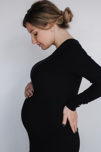 Elegant pregnant woman in beautiful black maternity drees put hand on belly in studio white