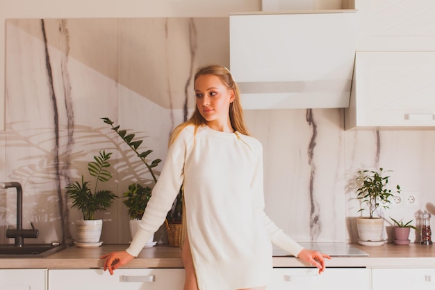 Elegant pretty girl in white long sleeve standing in modern kitchen feeling calm and relaxed looking away Thoughtful young woman daydreaming at home feeling comfortable beeing alone