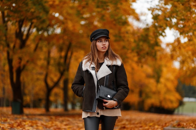 Elegant pretty young woman in a chic hat in a stylish jacket with a bag in the forest on a background of golden leaves. Cute girl walks in the park. Autumn collection of trendy women's clothing.